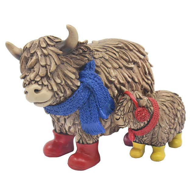 Decorative figurine of a highland cow with a blue scarf and red boots, standing next to a smaller cow figure with a red scarf and yellow boots on a white background.