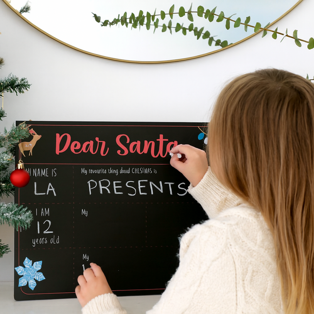 Person writing a letter to Santa Claus on a blackboard next to a Christmas tree.