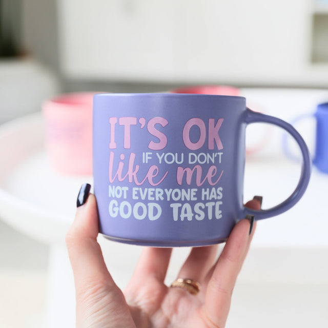 Person holding Splosh Cheeky Mug Good Taste, purple mug with humorous quote, perfect witty gift for friends or coworkers.