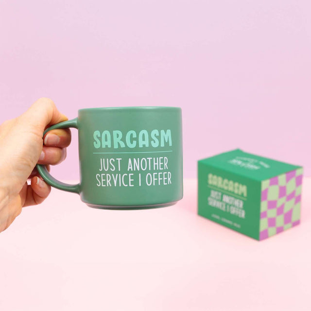 Person holding Splosh Cheeky Mug Sarcasm against pink background, humorous green mug with witty quote, perfect funny gift for him or her.